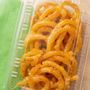 A closed, clear plastic clamshell container filled with crispy golden-orange spiral jalebi sweets resting on a light wooden background.